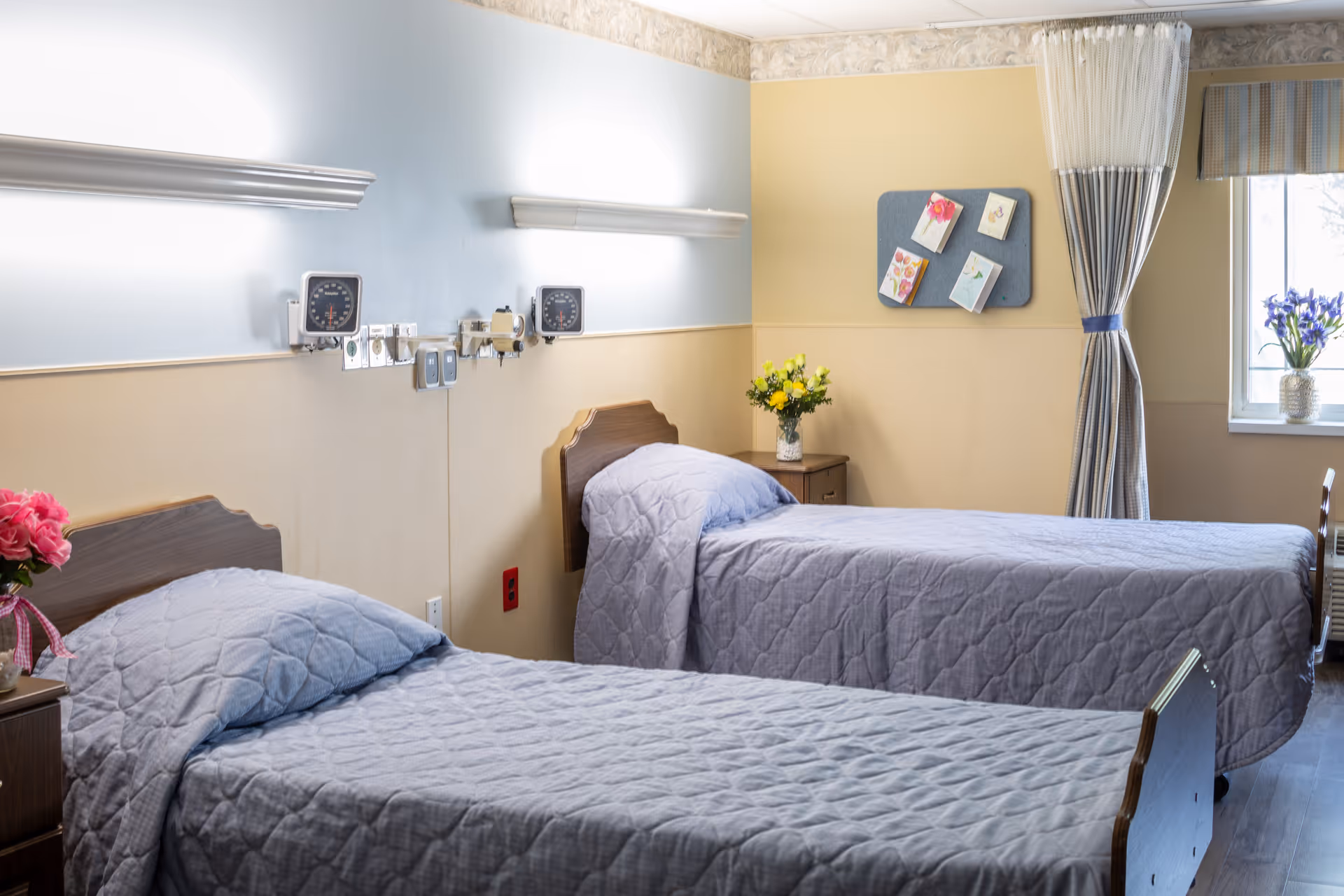 A bright and clean room with two single beds covered in light gray quilted bedspreads. Each bed has a wooden headboard and is accompanied by a small wooden nightstand with a vase of flowers. The walls are painted in two tones, light blue on the upper half and beige on the lower half. Medical equipment is mounted on the wall above the beds. A window with striped curtains and a vase of purple flowers is visible on the right side, and a bulletin board with cards is mounted on the far wall.