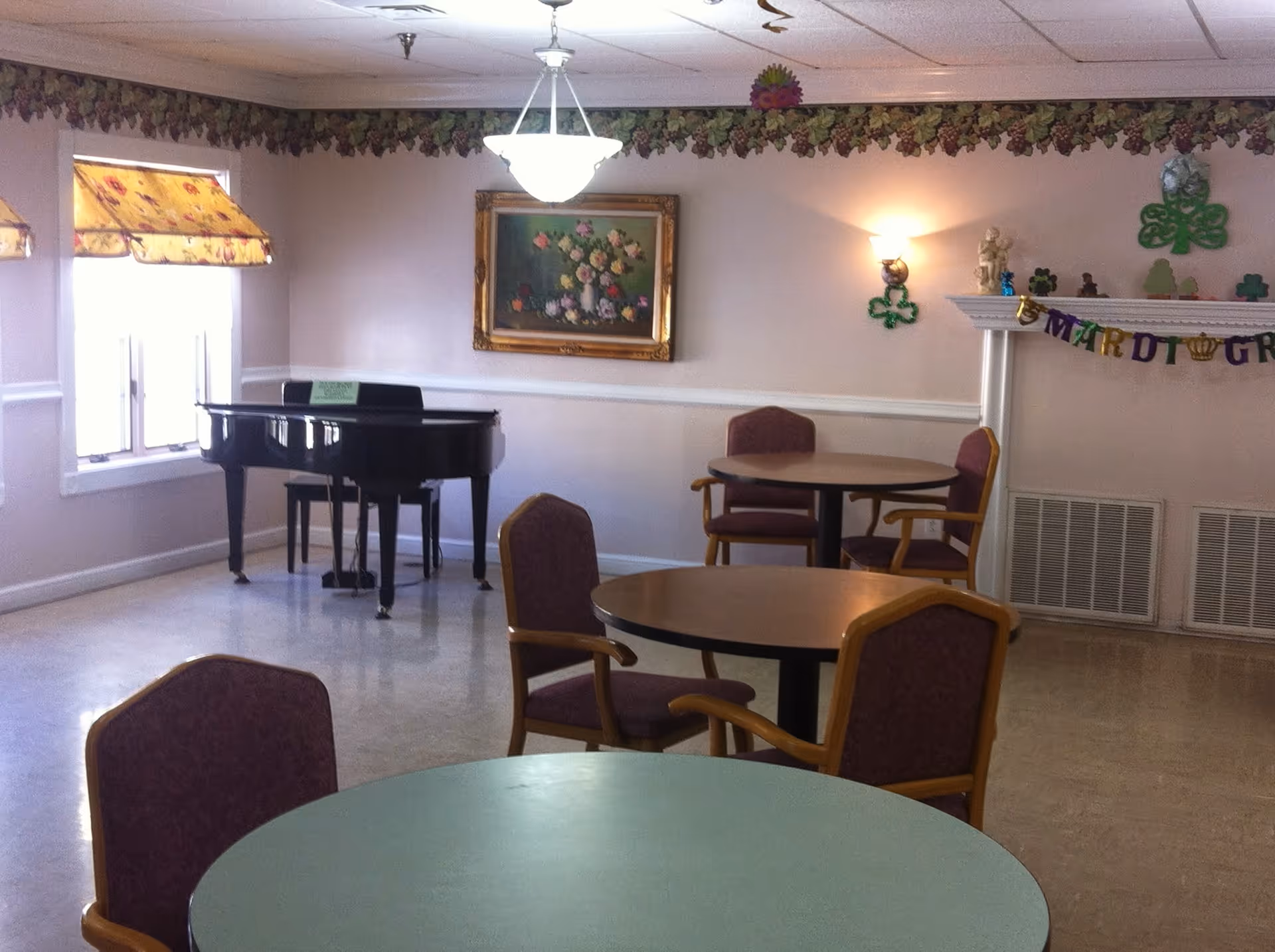 A common room with round tables and chairs, a black grand piano near two windows with floral valances, a floral painting on the wall, and a fireplace decorated with Mardi Gras themed items and shamrocks.