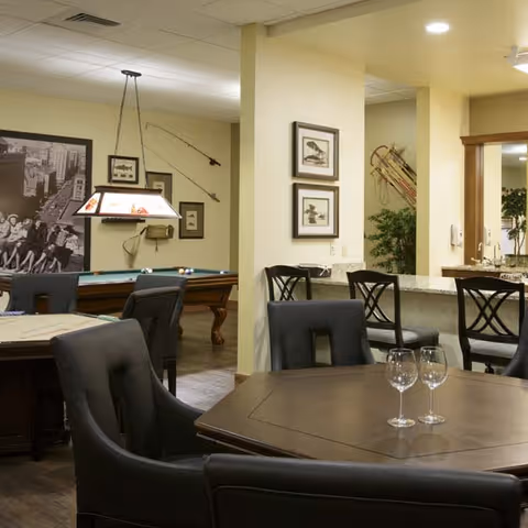 Interior view of a senior living facility common area featuring a pool table with a hanging light fixture above it, a dining table with four wine glasses, cushioned chairs, a bar counter with stools, framed artwork on the walls, and decorative plants.