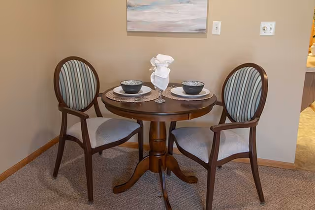 A small round wooden dining table set for two with two wooden chairs featuring striped upholstery. The table has two place settings with bowls, plates, and a glass with a folded white napkin. The setting is indoors with beige walls and carpeted floor.
