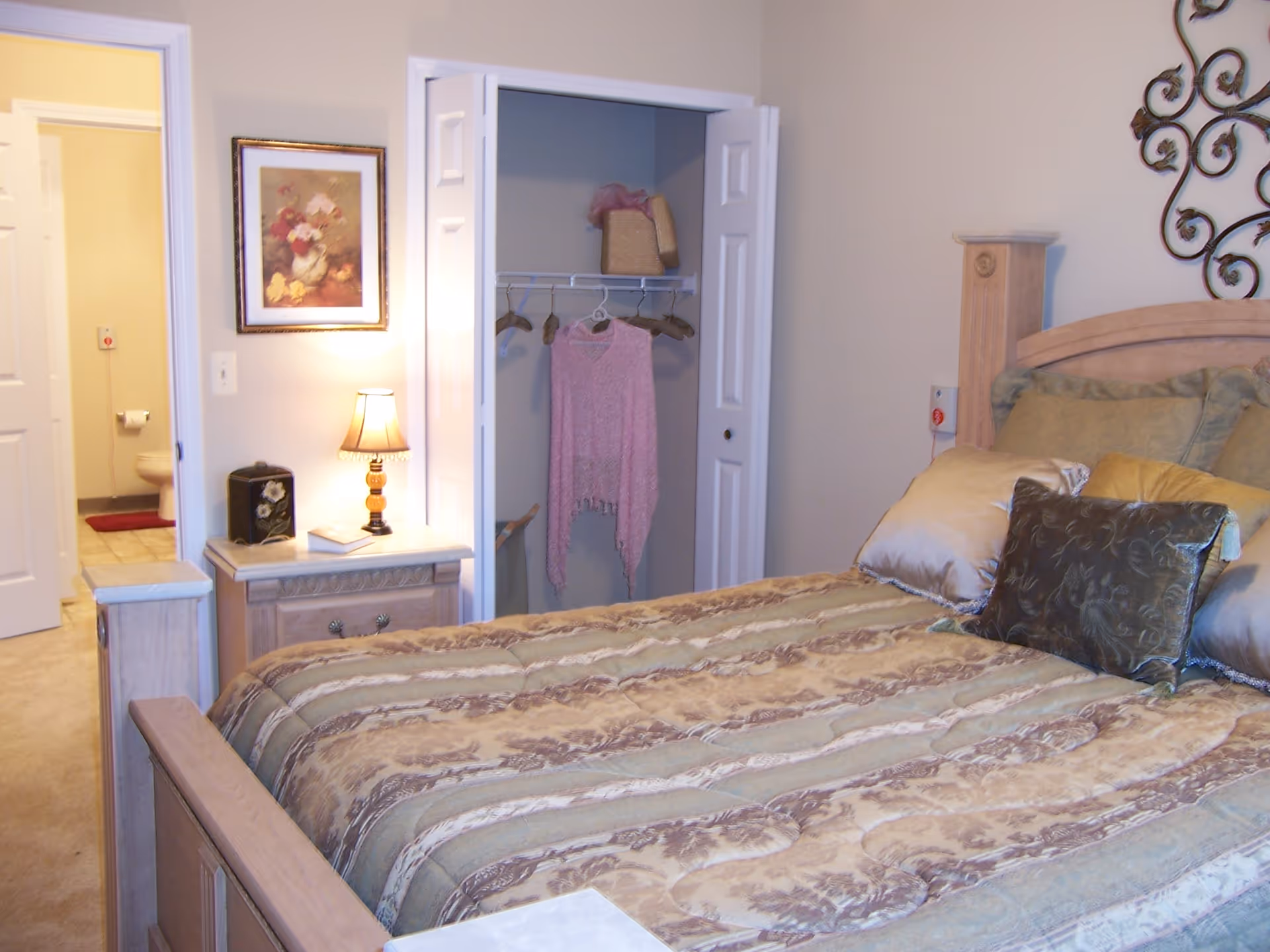 A furnished bedroom with a large bed, decorative pillows, a nightstand with a lamp, and an open closet and doorway to a bathroom.