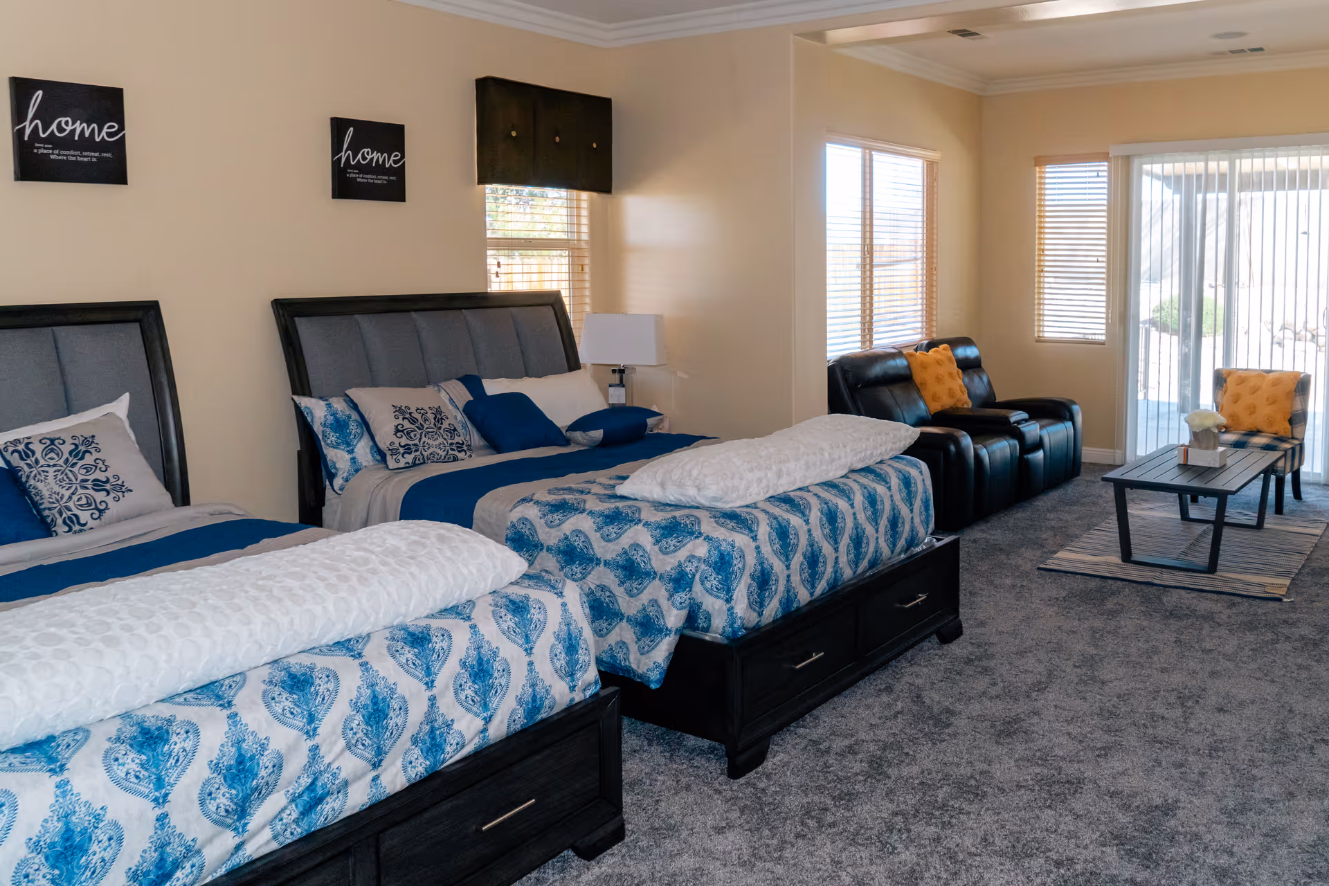 A spacious room with two beds featuring blue and white patterned bedding, a black leather loveseat with orange pillows, a matching chair, and a coffee table on a rug. The room has beige walls, carpeted floor, windows with blinds, and sliding glass doors leading outside. Two wall decorations with the word 'home' are hung above the beds.