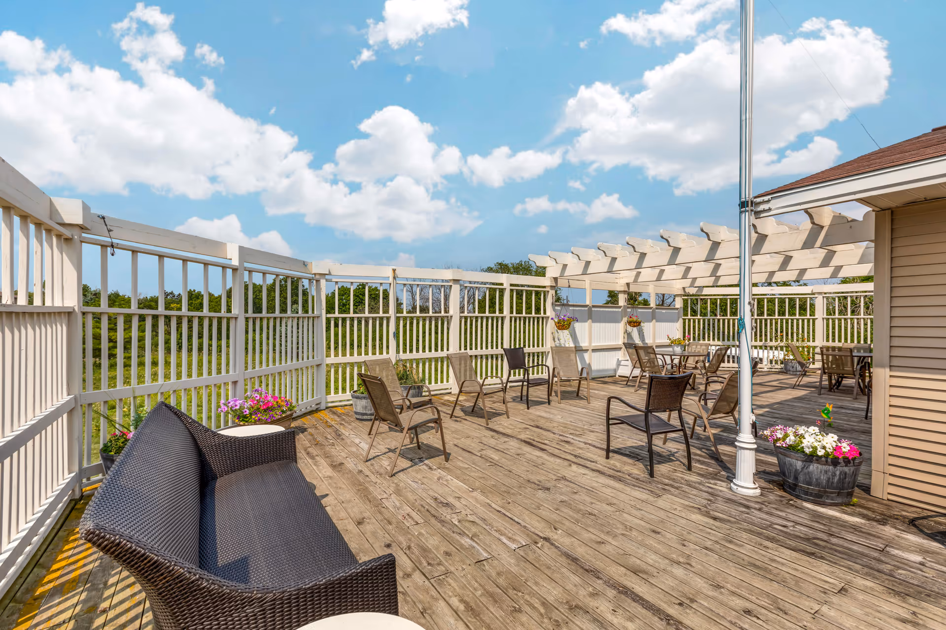 Wooden rooftop deck with chairs, a pergola, potted flowers and a wicker bench under a blue sky.