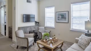 A cozy living room with two beige armchairs, a wooden coffee table with decorative items, a patterned rug, a wall-mounted flat-screen TV, and two windows with blinds. There is a framed picture on the wall and a table lamp on a side table.