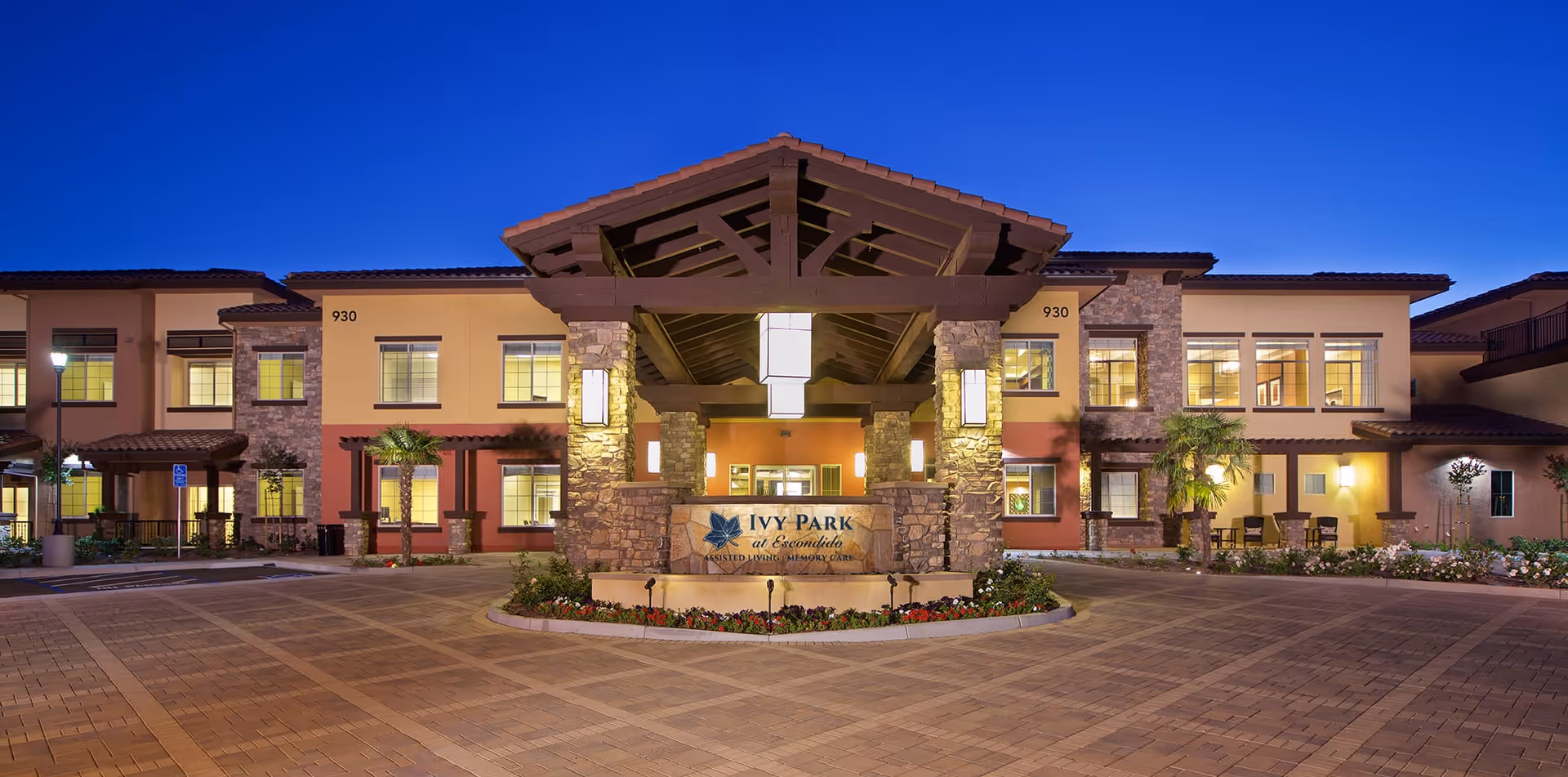 Front exterior view of Ivy Park at Escondido assisted living and memory care facility at dusk, showing a large entrance with stone pillars, a covered driveway, and well-lit windows.