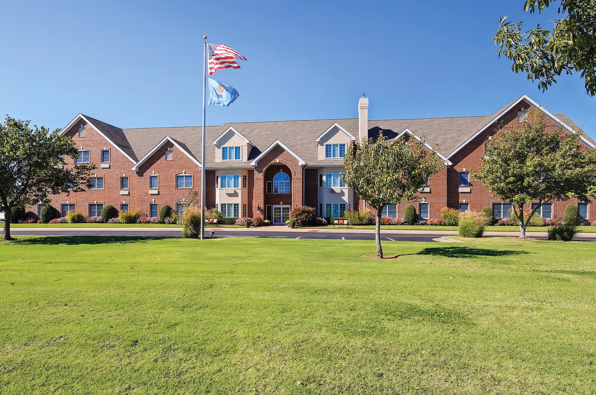 Front exterior view of a large brick assisted living facility building with multiple windows and a central entrance. Two flagpoles stand in front of the building, flying the American flag and a state flag. The building is surrounded by a well-maintained green lawn and several trees under a clear blue sky.