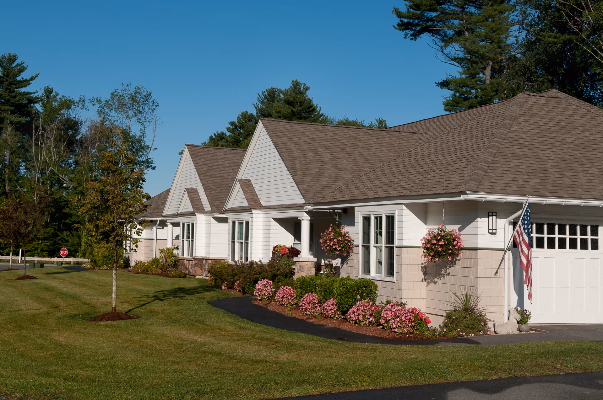 Front exterior of single-story cottage-style senior living units with landscaped lawn, hanging flower baskets, and an American flag.