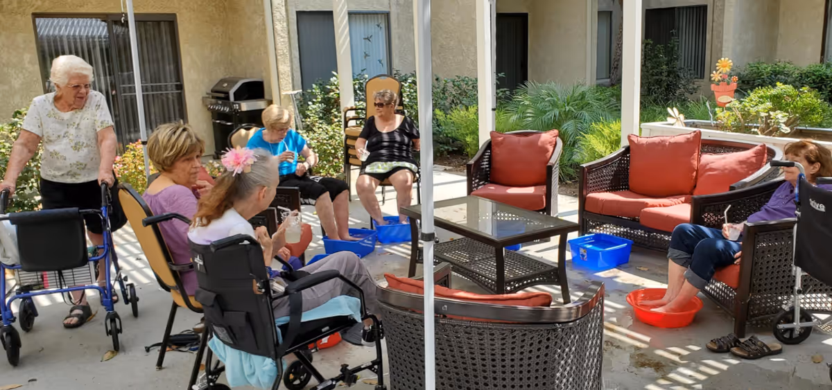 A group of elderly women sitting outdoors in a shaded patio area at a senior living facility. Some women have their feet soaking in blue and red plastic tubs filled with water. One woman is standing with a walker, and others are seated on chairs and cushioned wicker sofas around a glass-top coffee table. The area is surrounded by plants and windows of the building.
