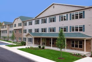 Three-story beige apartment building with a green roof, multiple windows, a covered ground-floor porch, and landscaped lawn and sidewalk.