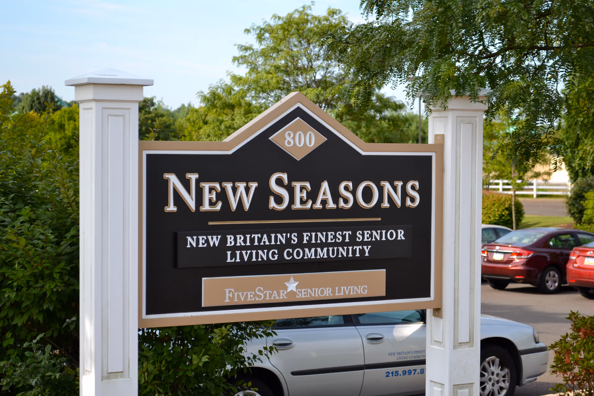 A large outdoor sign for New Seasons, New Britain's finest senior living community, with the Five Star Senior Living logo. The sign is mounted on white posts with greenery and parked cars visible in the background.