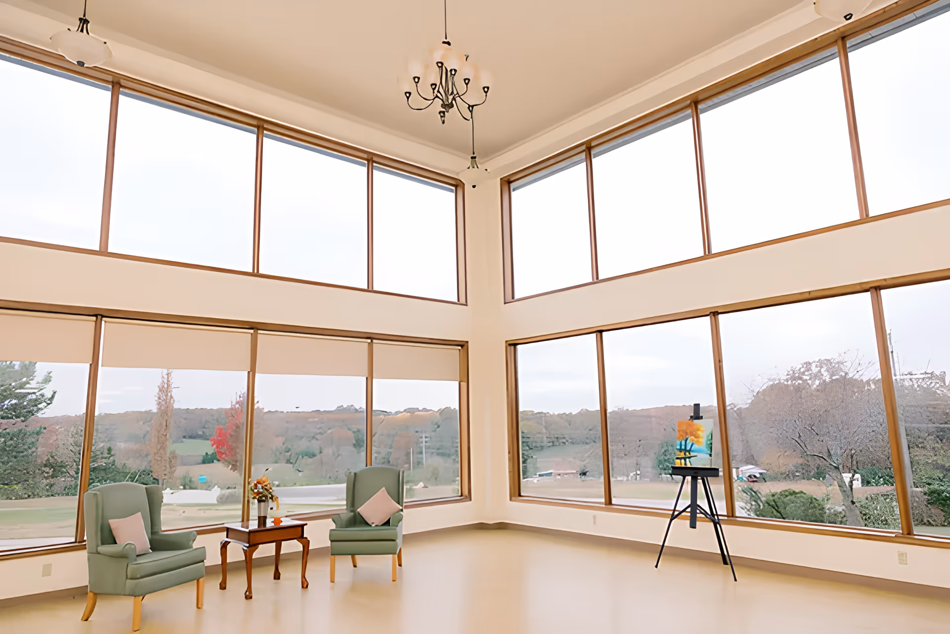 Bright two-story common sitting area with floor-to-ceiling windows, two green armchairs and a small table, and an easel overlooking a landscaped view.