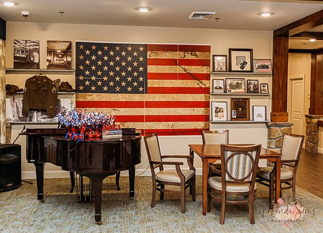 Interior room with a large American flag artwork on the wall, a black grand piano decorated with red, white, and blue floral arrangements, and a wooden table surrounded by four chairs. The walls display framed photographs and military memorabilia, including a military jacket. The room has a carpeted floor and warm lighting.