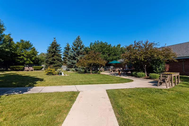 Outdoor garden area with green grass, trees, and shrubs under a clear blue sky. There are paved walkways, wooden benches, and a patio area with tables and an umbrella near a brick building.
