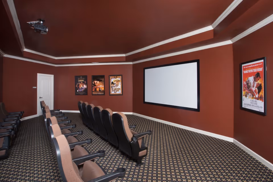 Interior view of a small movie theater room with rows of cushioned seats facing a large blank projection screen. The walls are painted dark red and decorated with framed movie posters. The ceiling has a projector mounted.