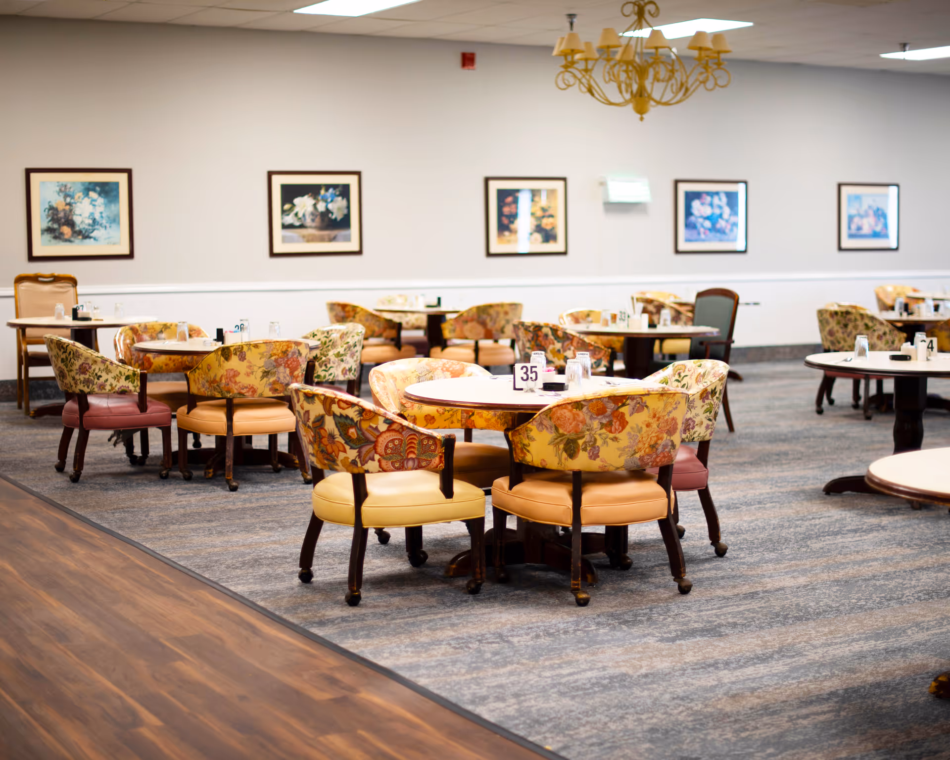 A dining room in a senior living facility with multiple round tables surrounded by floral-patterned chairs. The room has a carpeted floor with a wooden border, framed floral artwork on the walls, and a chandelier hanging from the ceiling.