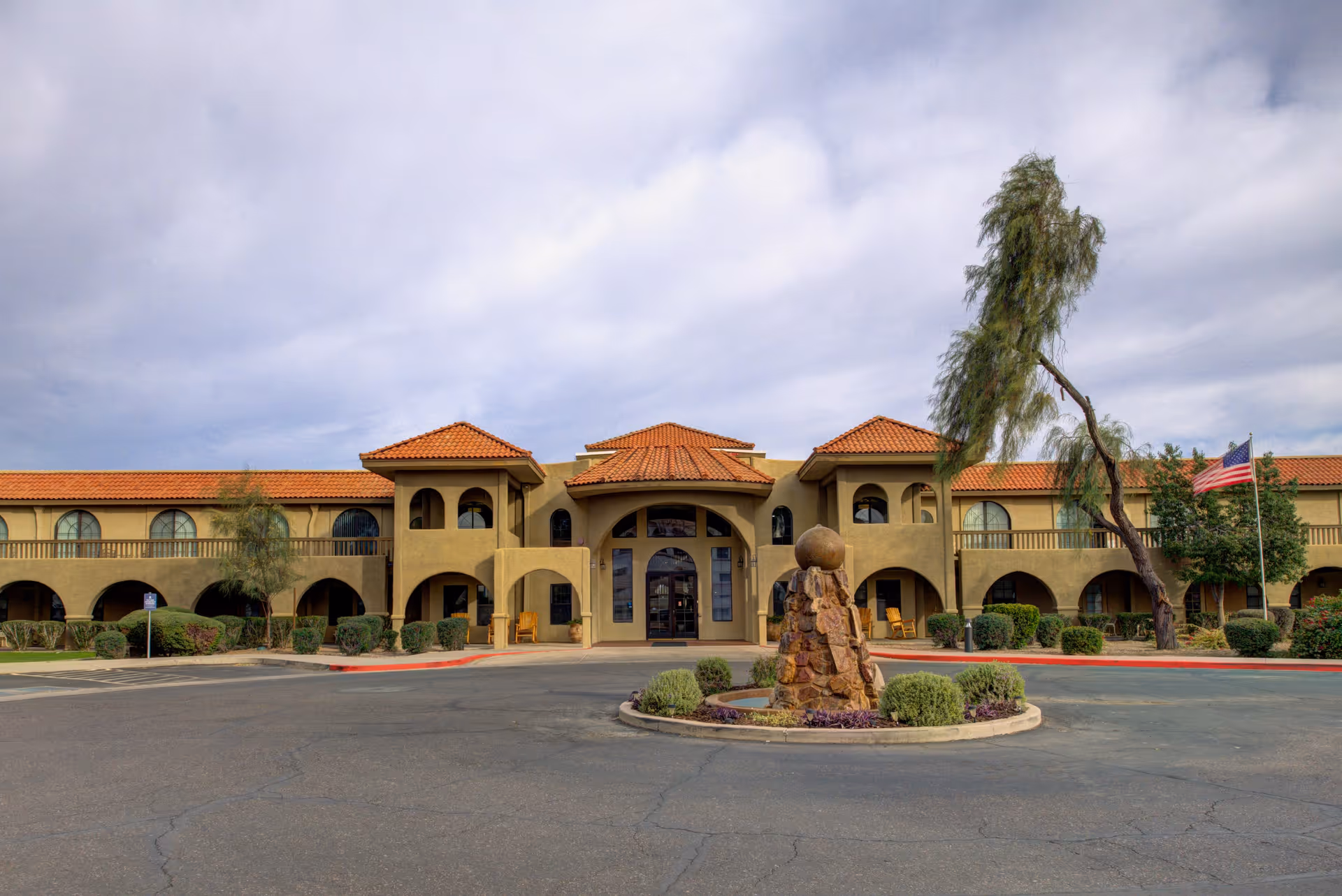 Front exterior view of The Garnet of Casa Grande facility with a two-story building featuring arched windows and doorways, a red tile roof, a circular driveway with a stone fountain centerpiece, some landscaping with bushes and trees, and an American flag on the right side.