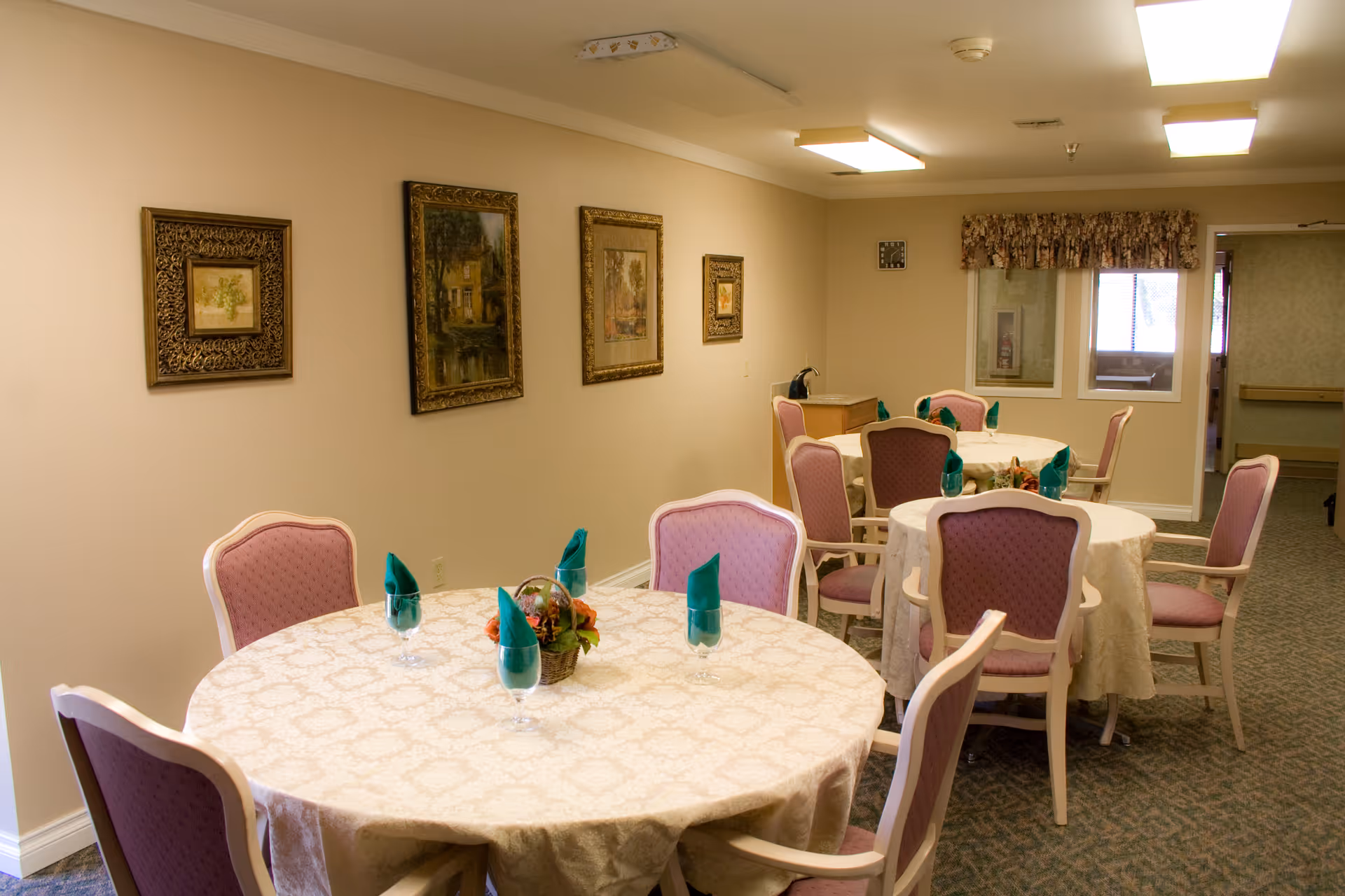 A dining room with round tables covered with beige tablecloths, each set with four chairs upholstered in pink fabric. Each table has glasses with green folded napkins and a small floral centerpiece. The walls are decorated with framed paintings, and there is a window with a floral valance in the background.