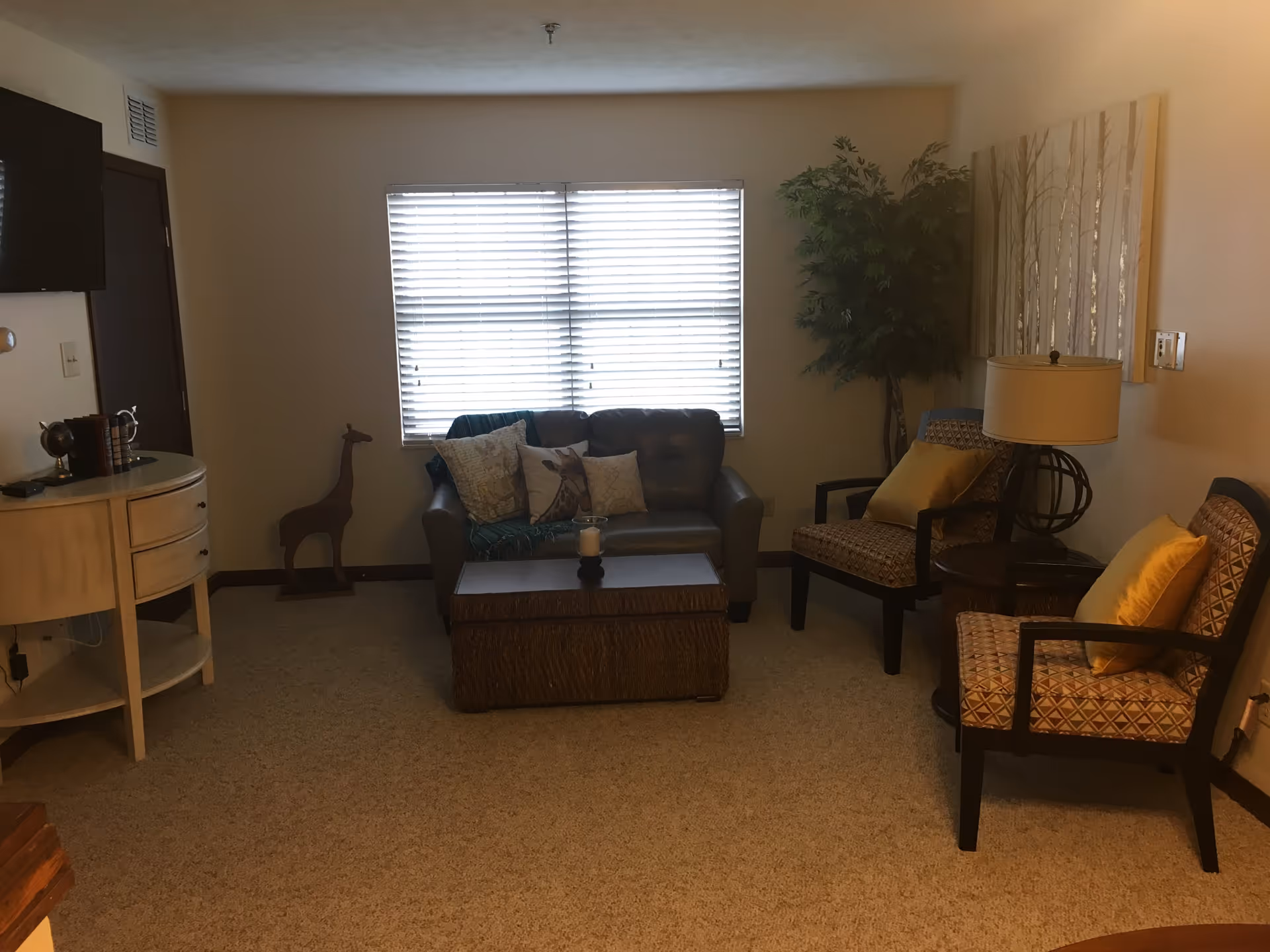 A cozy living room with a small brown leather loveseat adorned with decorative pillows, a wooden coffee table with a candle on top, two patterned armchairs with yellow cushions, a round side table with a lamp, a large potted plant in the corner, a window with blinds, a wall-mounted TV, and a small wooden giraffe figurine near a white console table.