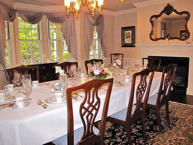 A formal dining room with a long table covered with a white tablecloth, set with plates, cups, glasses, and silverware. There is a floral centerpiece on the table. The room has large windows with draped curtains, a chandelier hanging from the ceiling, a fireplace with a decorative mirror above it, and framed artwork on the wall.