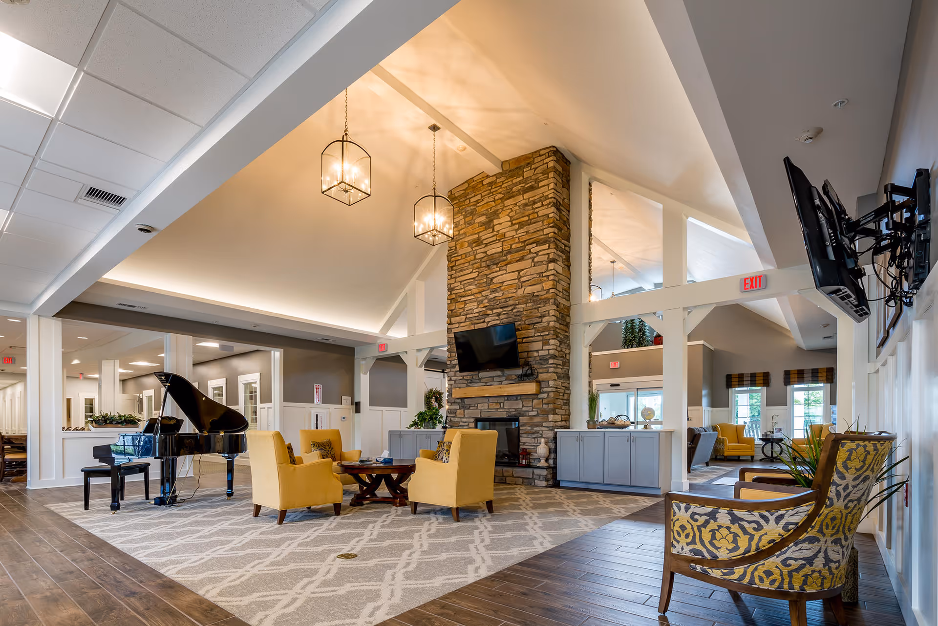 Spacious senior living facility common area with a high vaulted ceiling, stone fireplace, mounted TV, and modern pendant lights. The room features a grand piano, yellow armchairs around a wooden coffee table on a patterned rug, and additional seating with patterned chairs. Large windows and glass doors allow natural light to fill the space.