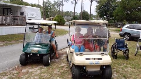 Two golf carts with people seated inside are parked on a dirt path in an outdoor area with trees, a wheelchair, and vehicles in the background.