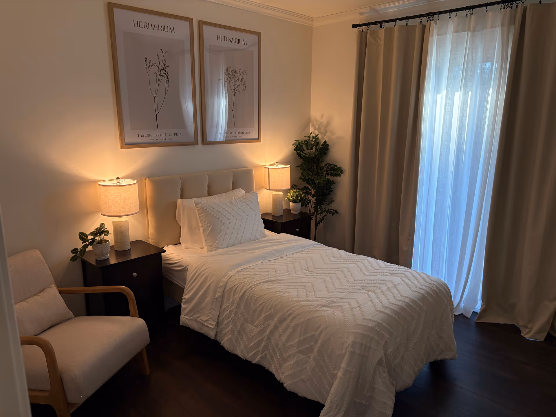 A cozy bedroom with a single bed covered in a white textured blanket and matching pillow. There are two bedside tables with lamps and small potted plants on each side of the bed. Two framed botanical prints hang above the bed. A beige armchair is positioned to the left, and a large window with sheer white curtains and beige drapes is on the right side of the room. The floor is dark wood.