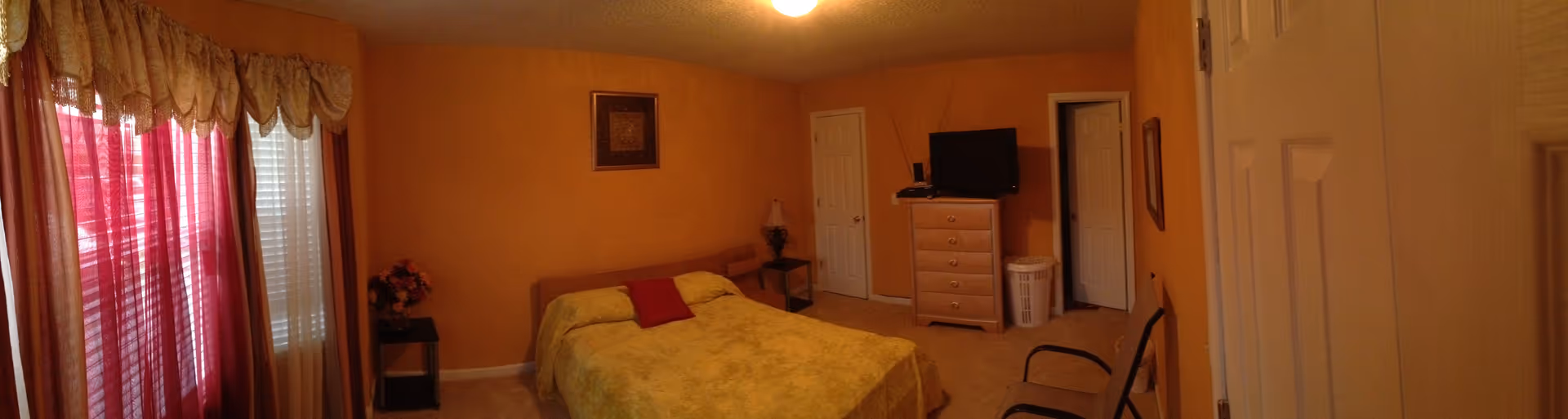A bedroom with orange walls, a bed covered with a yellow bedspread and a red pillow, two small side tables with lamps, a dresser with a television on top, a laundry basket, a chair, and windows with red and beige curtains.