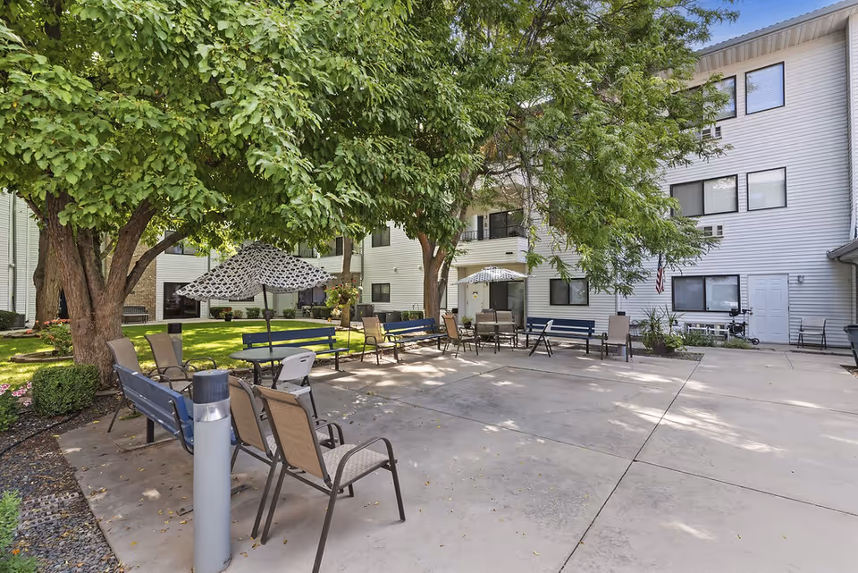 Outdoor courtyard area at Karcher Senior Living with several benches and chairs arranged around tables with umbrellas, surrounded by trees and adjacent to a white multi-story building.