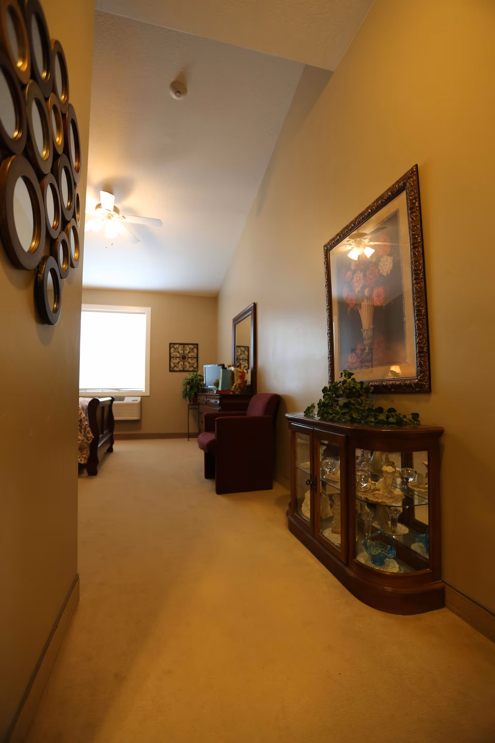 Carpeted interior corridor of a care suite showing a glass display cabinet, framed art, an armchair and a bed by a window.