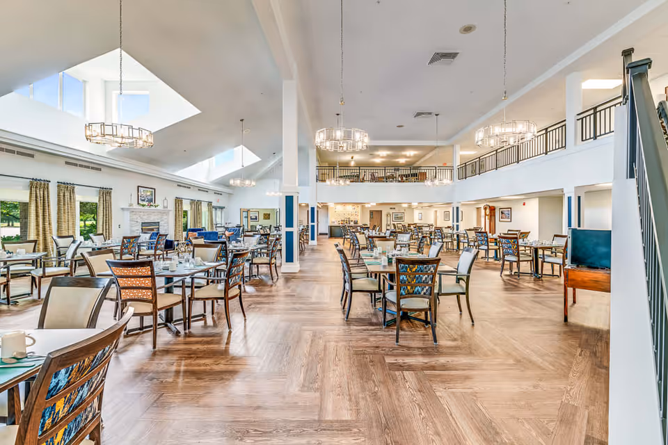 A spacious and bright dining room in a senior living facility with multiple tables and chairs arranged neatly. The room features large windows with curtains, a fireplace, modern chandeliers hanging from the ceiling, and a mezzanine level with railings overlooking the dining area. The floor is wooden, and the overall atmosphere is clean and inviting.