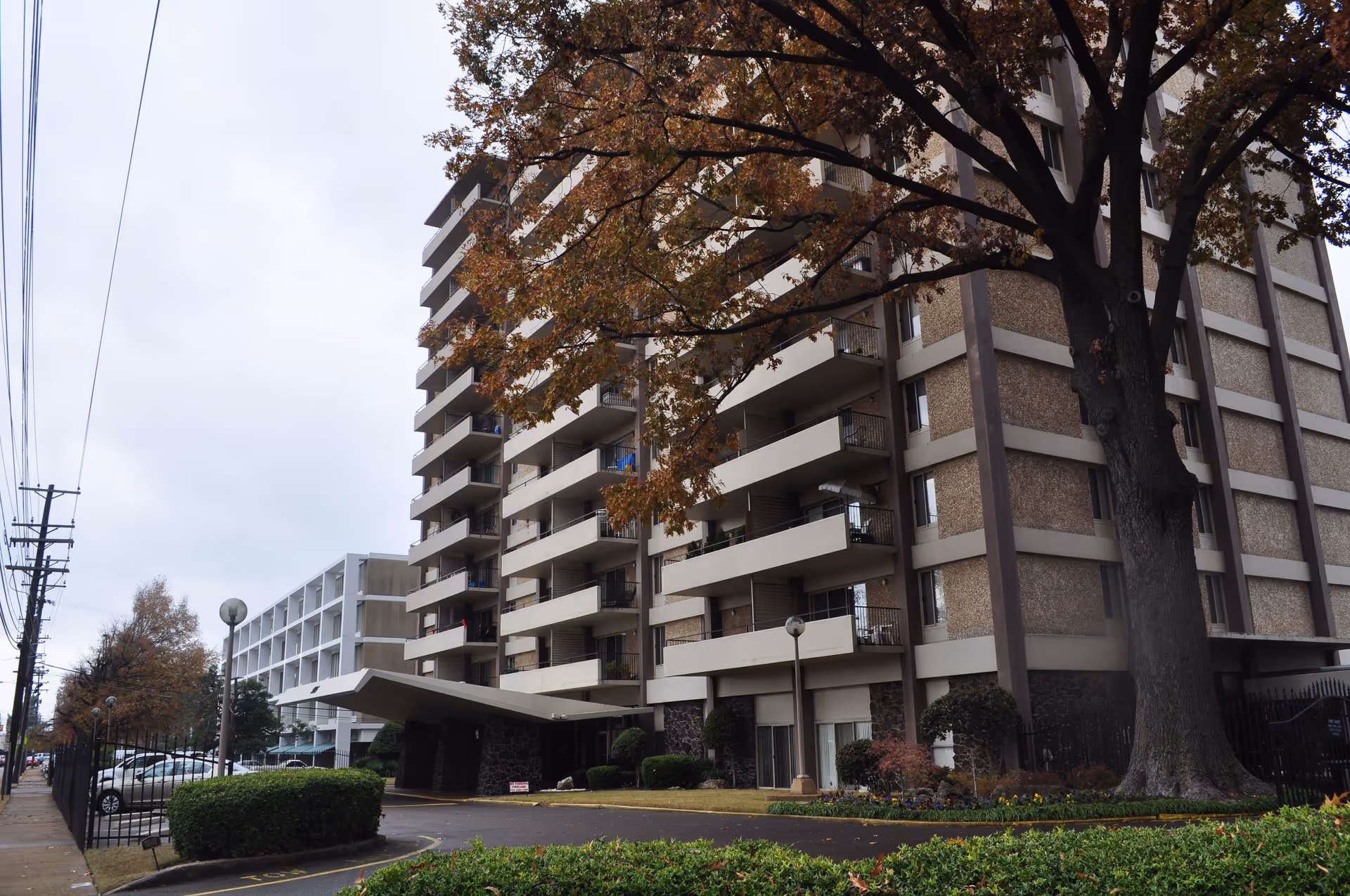 Exterior view of a multi-story residential building with balconies, surrounded by trees and bushes. A driveway leads to the entrance, and there are parked cars visible along the side. The sky is overcast.
