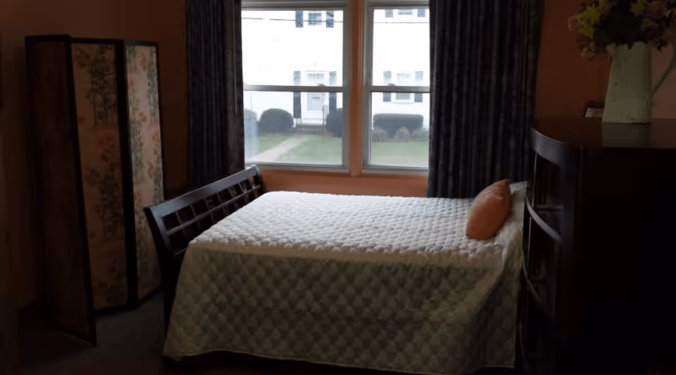 A bedroom with a bed covered in a white quilt and an orange pillow, positioned in front of two large windows with dark curtains. To the left of the bed is a decorative folding screen with floral patterns, and to the right is a dark wooden dresser with a vase of flowers on top.