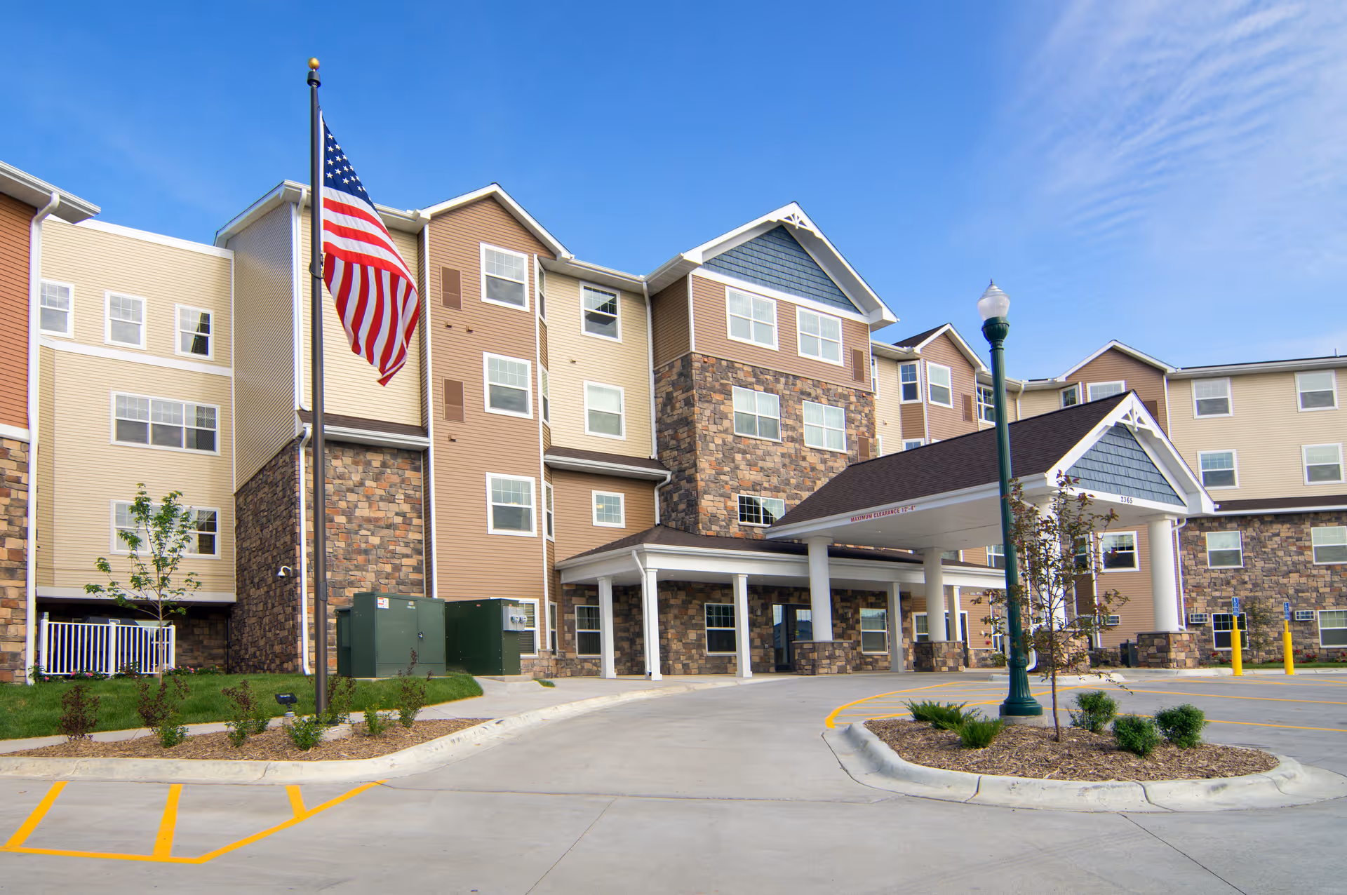 Front exterior of a multi-story senior living building with a covered entrance, driveway, and an American flag on a flagpole.