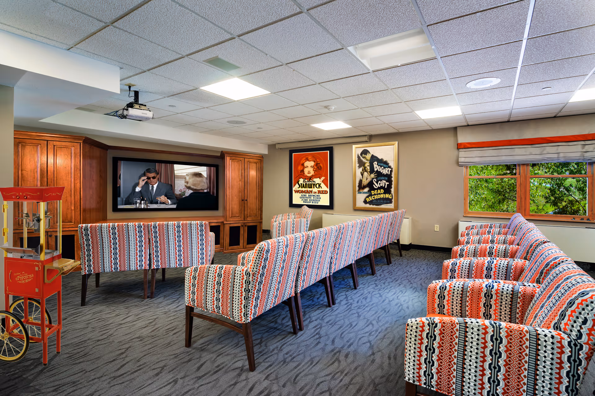 A cozy media room with patterned upholstered chairs arranged in rows facing a large flat-screen TV mounted on a wooden cabinet. The TV is playing a black-and-white movie. There are two vintage movie posters on the wall and a red popcorn machine on the left side of the room. A window with a view of green trees is on the right side.