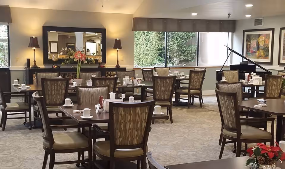 A dining room with multiple tables and chairs arranged neatly. Each table has white cups, napkins, and condiments. There are two lamps on a sideboard with a large mirror above it, a large window with a view of greenery, framed artwork on the walls, and a grand piano in the corner.