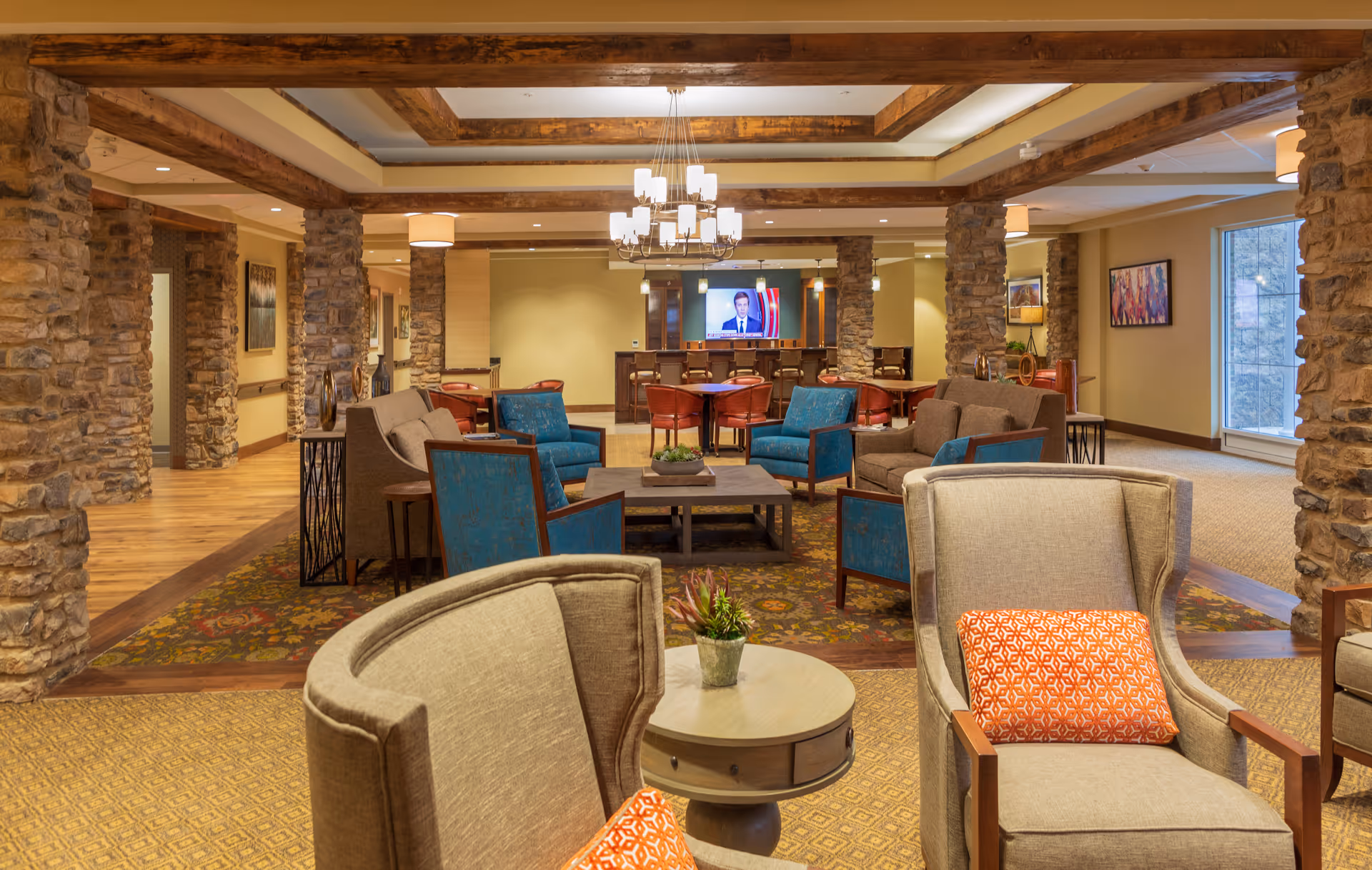 A spacious senior living community lounge area with stone pillars and wooden beams on the ceiling. The room features comfortable seating including beige armchairs with orange patterned pillows, blue armchairs, and brown sofas arranged around coffee tables. A chandelier hangs from the ceiling, and a television is mounted on the far wall above a bar area with stools. The room is warmly lit and decorated with framed artwork and plants.