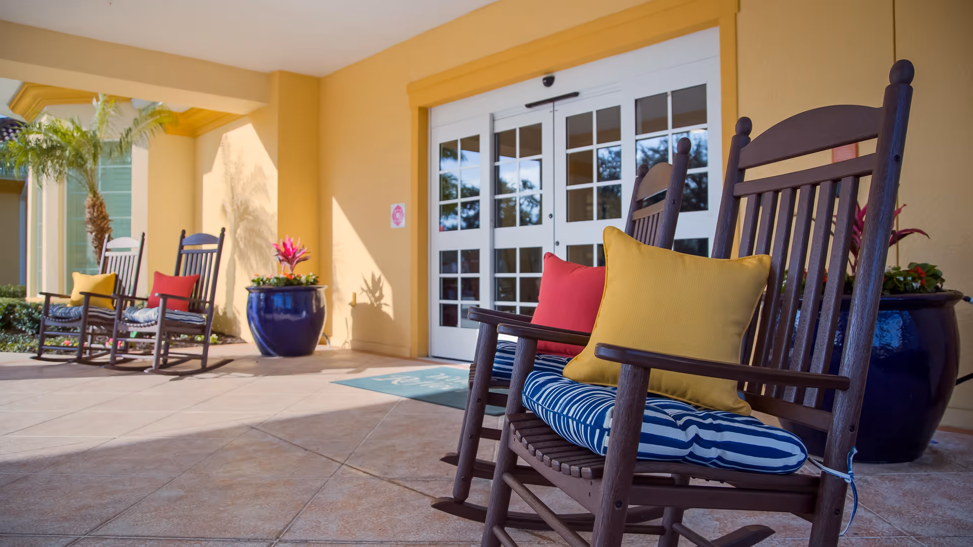 Outdoor patio area at Windsor Reflections Memory Care Of Lakewood Ranch with wooden rocking chairs featuring colorful cushions, large blue ceramic planters with plants, and a double glass door entrance under a yellow exterior wall.