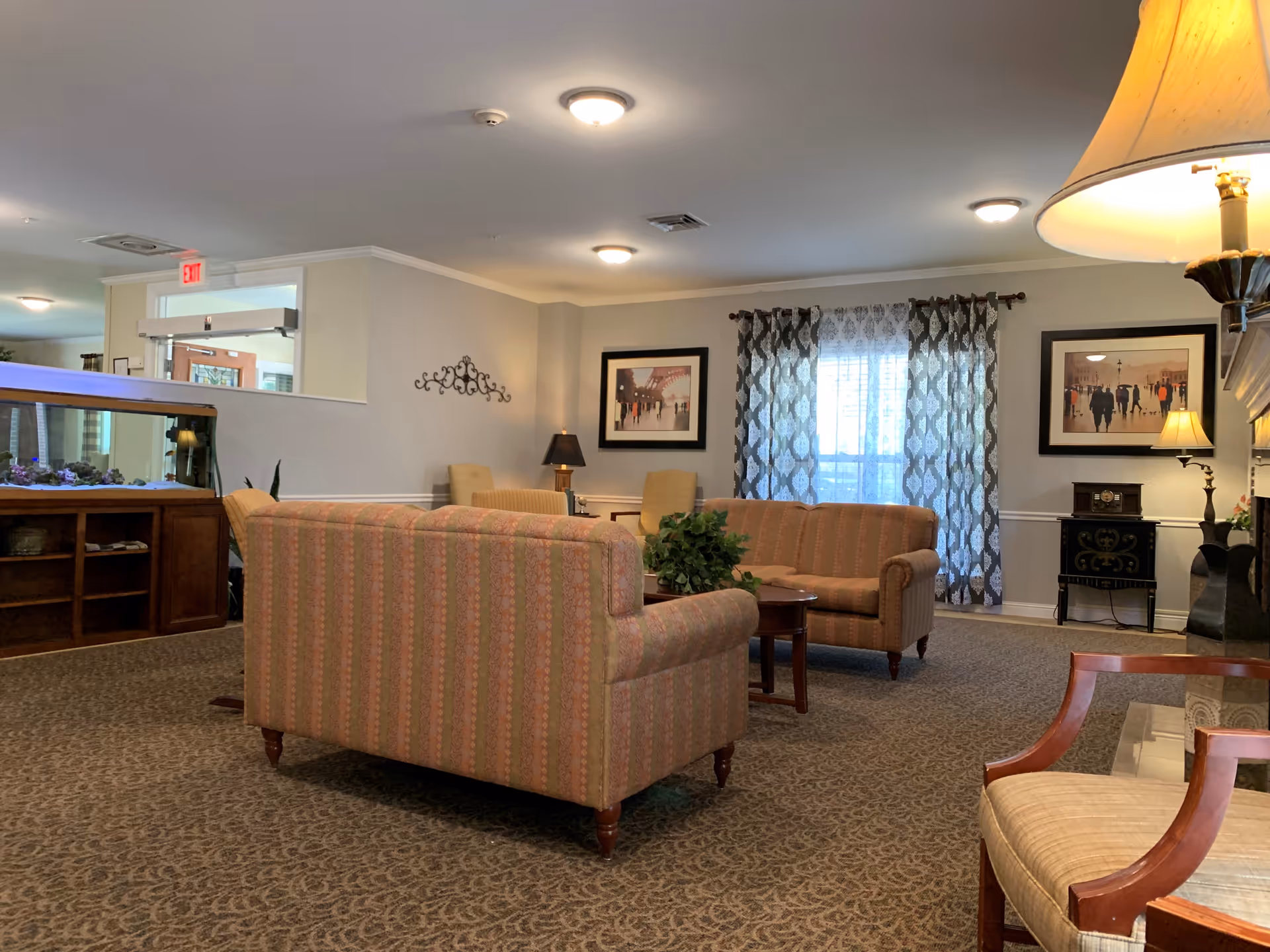 A cozy assisted-living common room with upholstered sofas, armchairs, a coffee table, framed artwork, and patterned curtains.