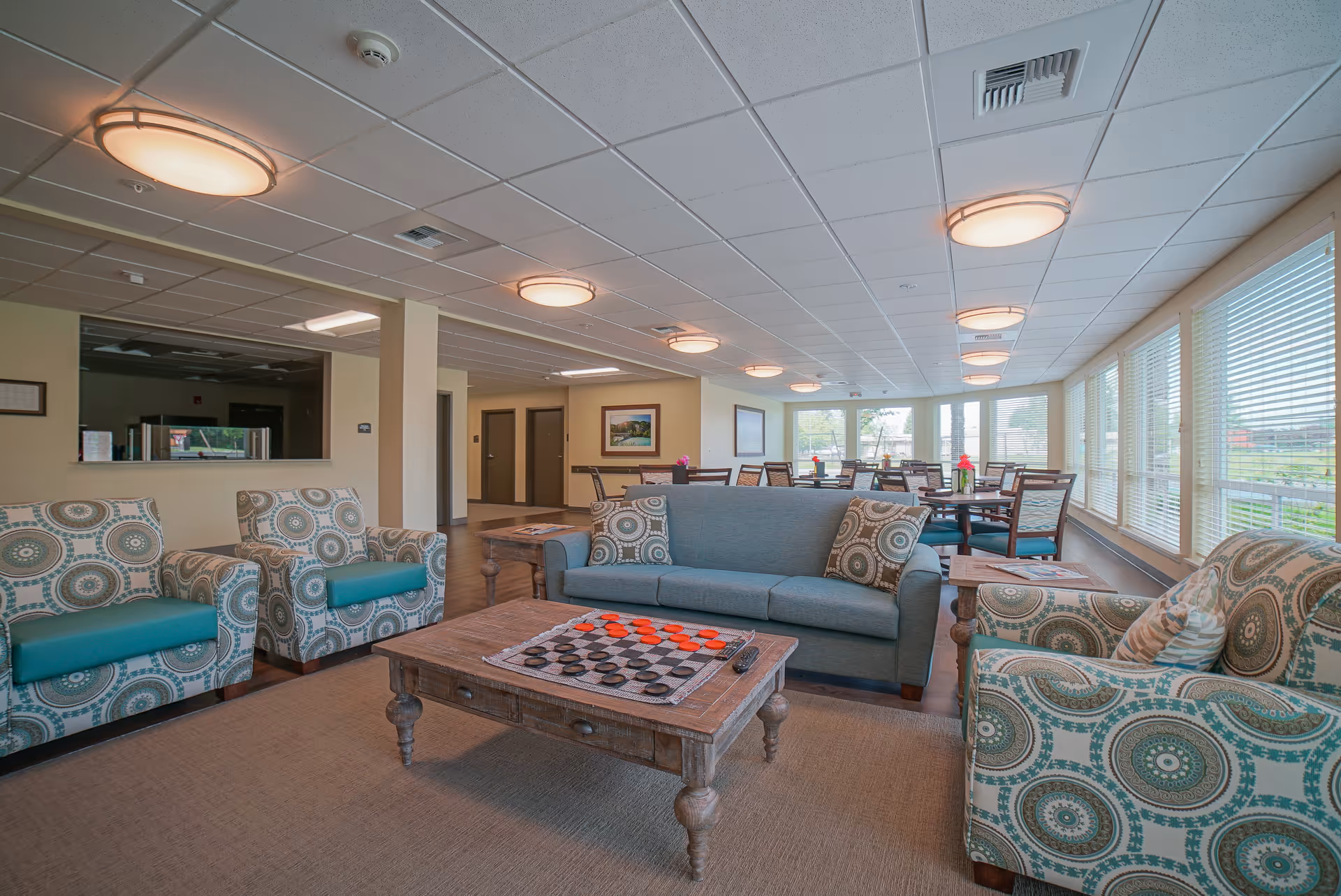 A spacious and well-lit common area in a senior living facility featuring a seating arrangement with a blue sofa and patterned armchairs around a wooden coffee table with a checkers game set up. In the background, there are multiple dining tables and chairs near large windows with blinds, allowing natural light to fill the room.