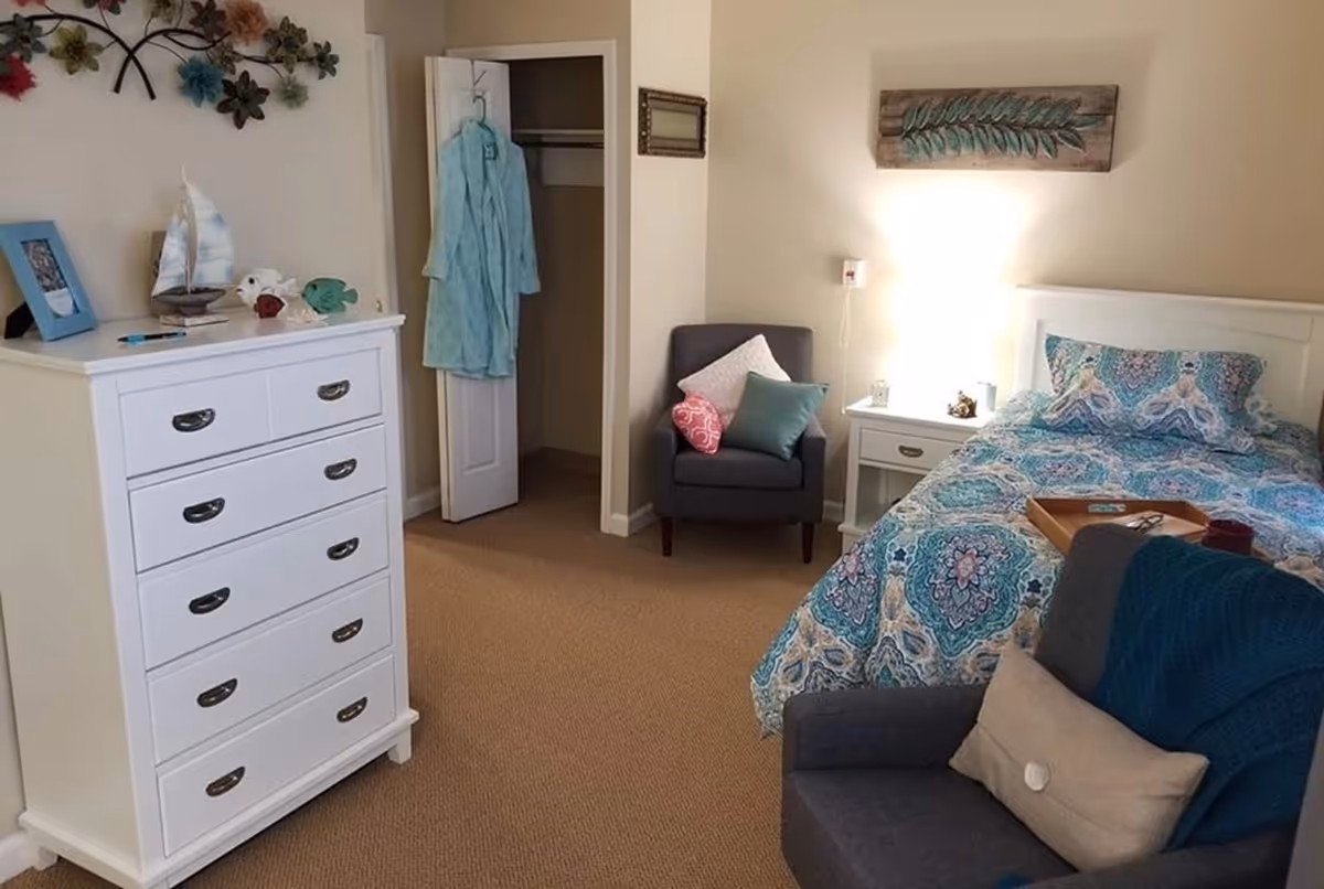 A cozy bedroom with a white dresser on the left, a small open closet with a blue robe hanging inside, a gray armchair with colorful pillows, a white nightstand with a lamp, and a bed with a blue patterned bedspread. There is a decorative wall piece above the bed and a second gray armchair with a beige pillow and blue throw in the foreground.