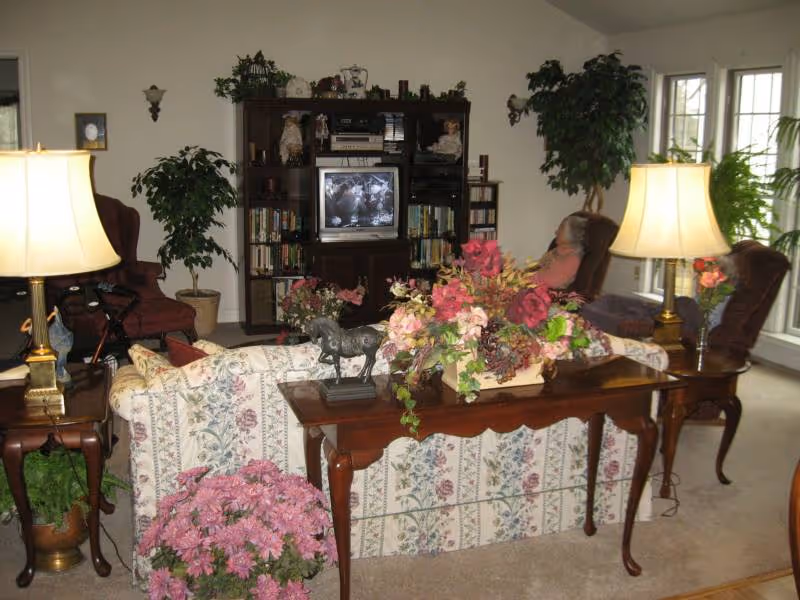 A cozy living room with floral patterned sofa, two armchairs, wooden side tables with lamps, a wooden console table with a floral arrangement and a horse figurine. There is a large entertainment center with a TV and bookshelves, and potted plants around the room. A person is seated in one of the armchairs near the windows.