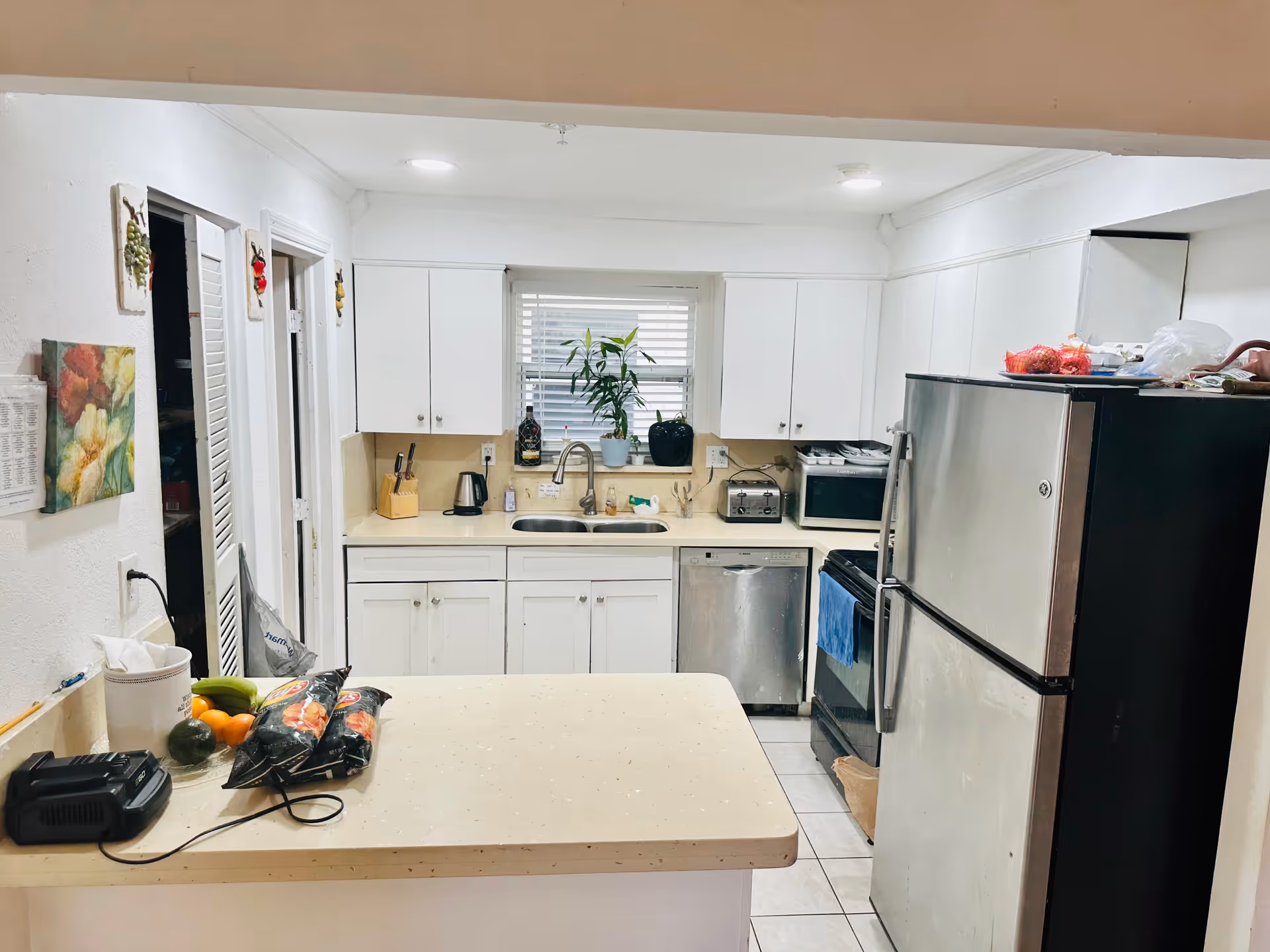 A clean kitchen with white cabinets and countertops. There is a double sink under a window with blinds, a stainless steel dishwasher, a stainless steel refrigerator, a black stove with a blue towel hanging on the handle, a microwave, a toaster, and various kitchen items including a knife block and a potted plant on the windowsill. On the counter in the foreground, there are some groceries including bananas, oranges, and packaged food items.
