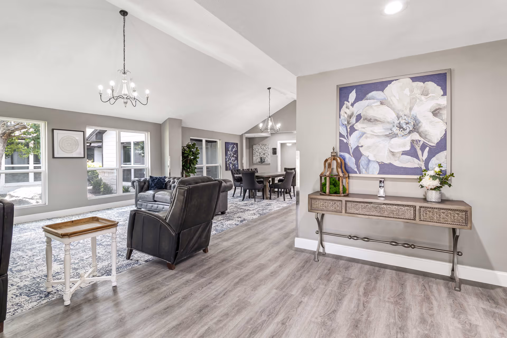 Bright open living area with leather armchairs, a console table topped with decor and floral artwork, and a dining area visible in the background.