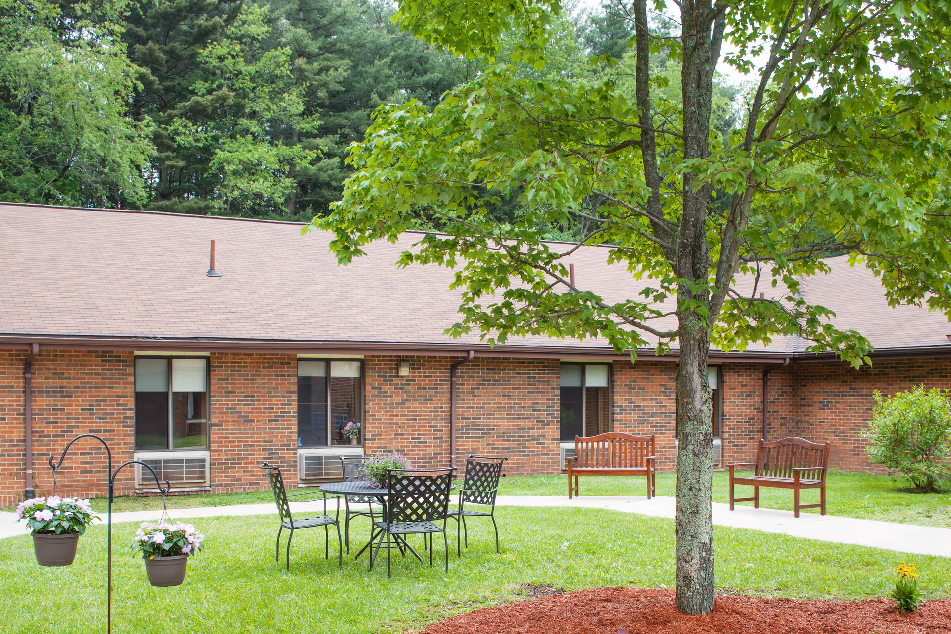 Courtyard outside a single-story brick senior living building featuring a tree, patio table and chairs, wooden benches, and hanging flower pots.