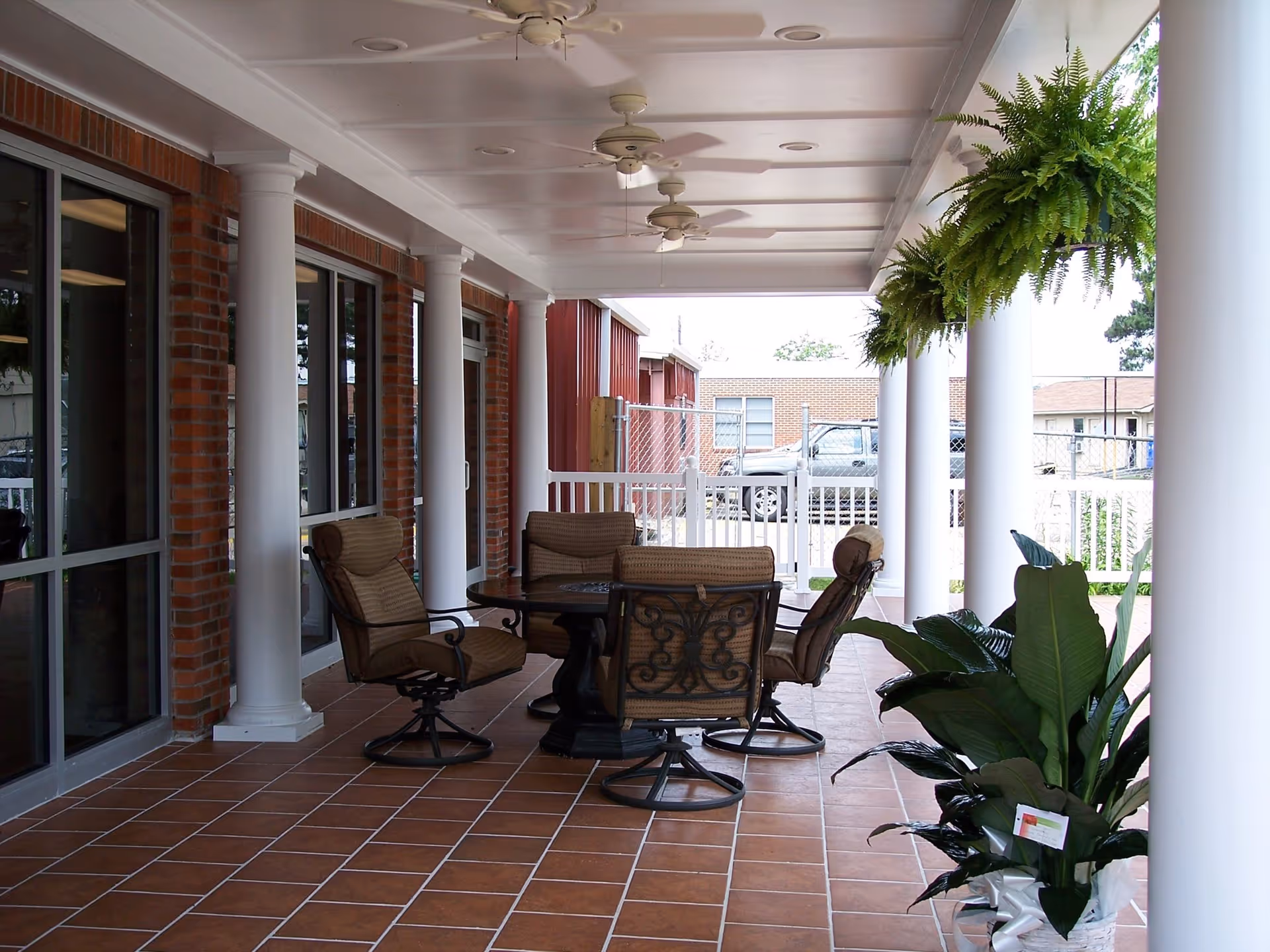 Covered outdoor patio area with a tiled floor, white columns, ceiling fans, and a round table surrounded by four cushioned chairs. There are hanging ferns and potted plants along the edge of the patio, with a view of a parking area and buildings in the background.