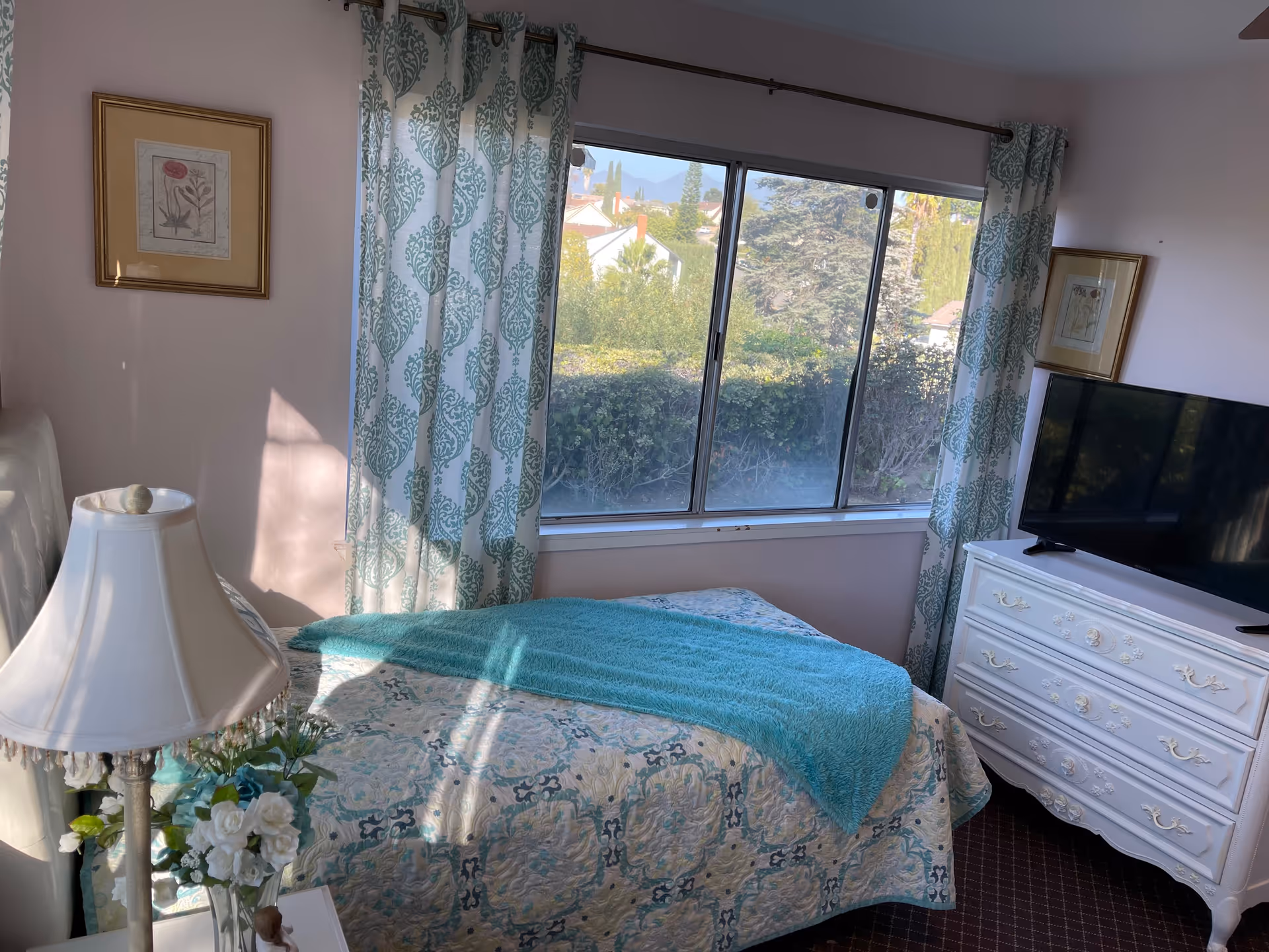 A cozy bedroom with a single bed covered in a patterned quilt and a teal throw blanket. Next to the bed is a white nightstand with a lamp and a vase of white flowers. The room has a large window with patterned curtains, letting in natural light and showing greenery outside. A white dresser with a flat-screen TV on top is positioned against the wall. Two framed floral pictures hang on the walls.