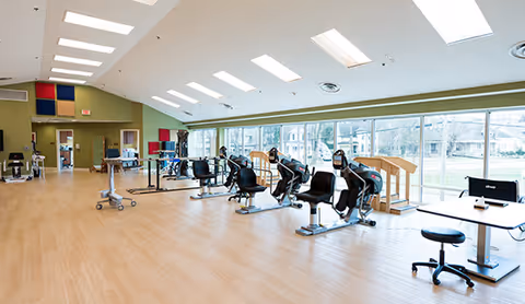 Spacious rehabilitation gym with stationary exercise bikes, other therapy equipment, and large windows overlooking a lawn.