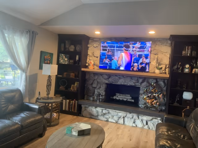 Cozy living room with stone fireplace, built-in shelves, and a wall-mounted TV in a senior facility.