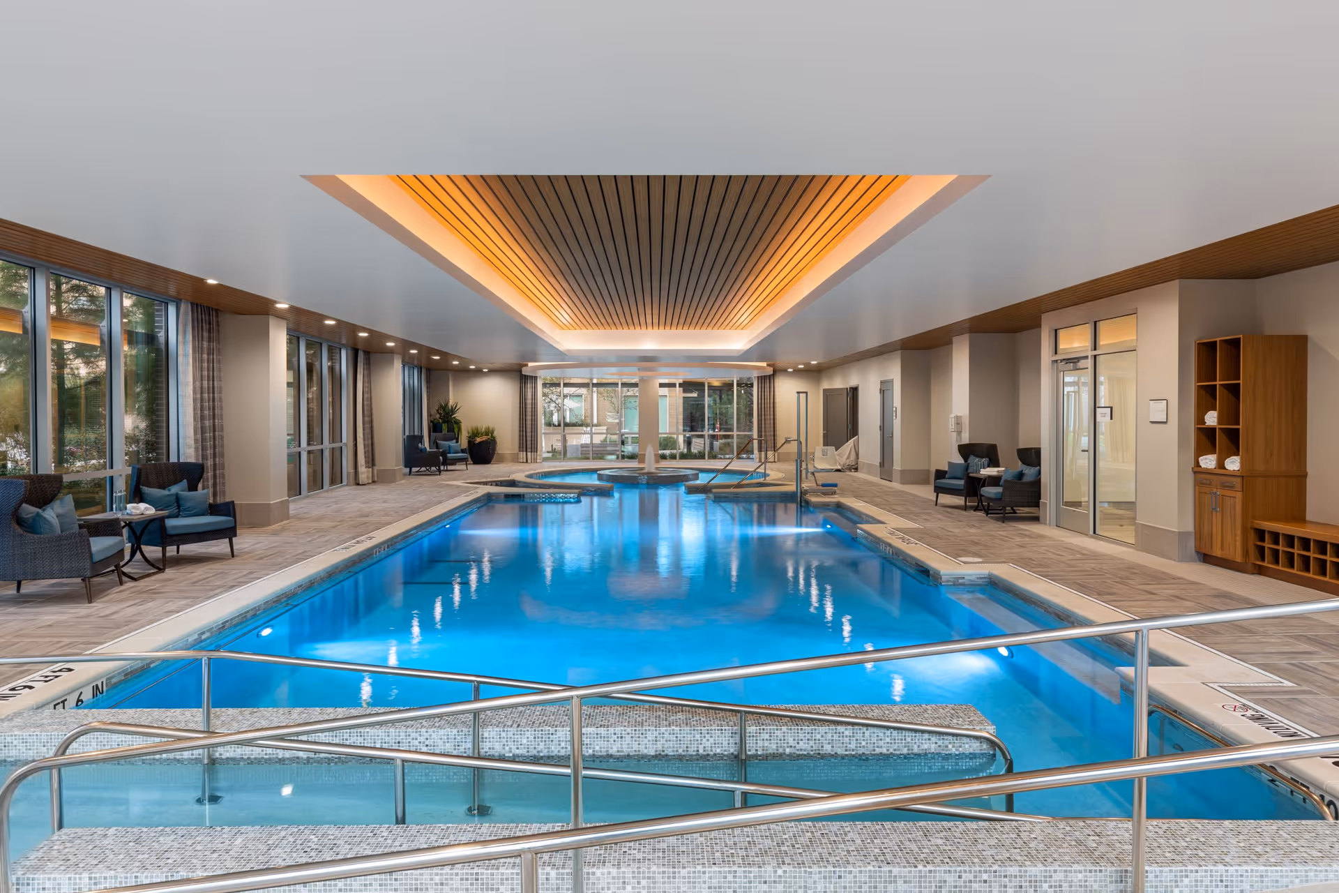 Indoor swimming pool area with clear blue water, surrounded by seating areas with chairs and small tables. The ceiling features recessed lighting and wooden paneling. Large windows line one side of the room, allowing natural light to enter. There are handrails and steps leading into the pool, and a hot tub is visible at the far end.