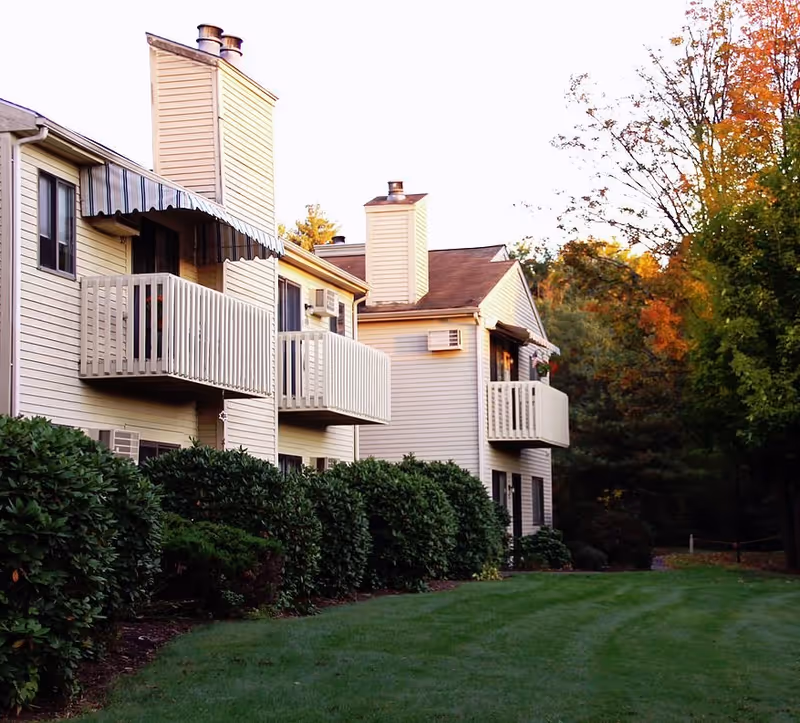 Exterior view of a residential building with beige siding, multiple balconies, and chimneys. The building is surrounded by green bushes and a well-maintained lawn, with trees showing autumn foliage in the background.